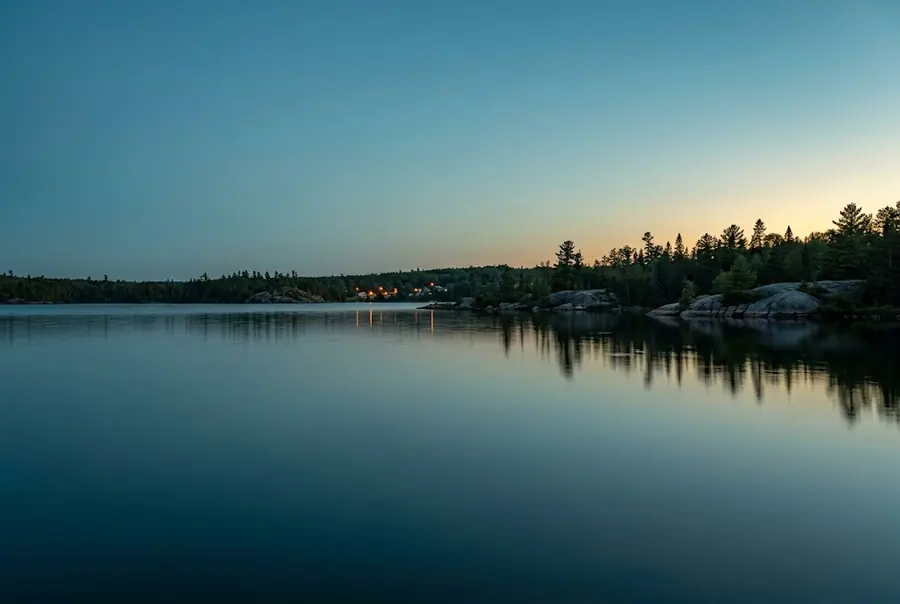 A northern lake at dusk, with a treeline and distant shoreline lights